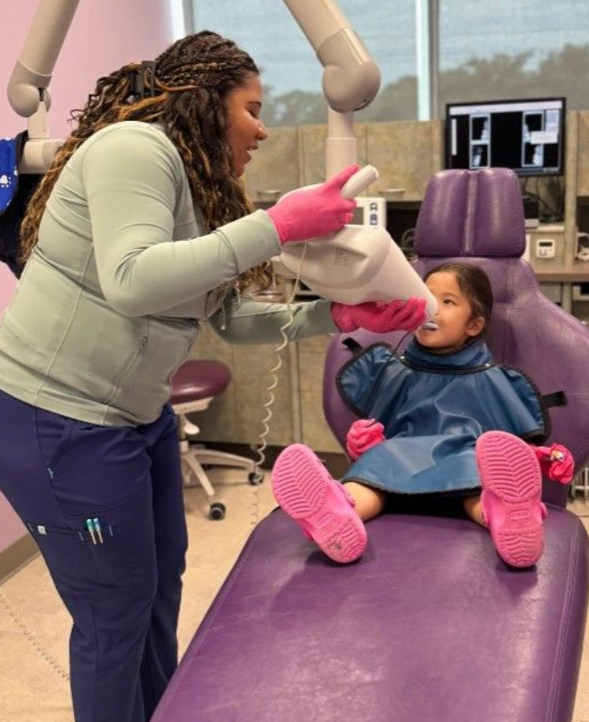 Dentist rinsing a child’s mouth in a purple dental chair, with colorful socks and a clinic monitor nearby