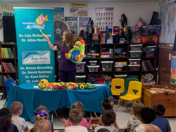 Children watch a woman presenting at a colorful table in a classroom or library setting.