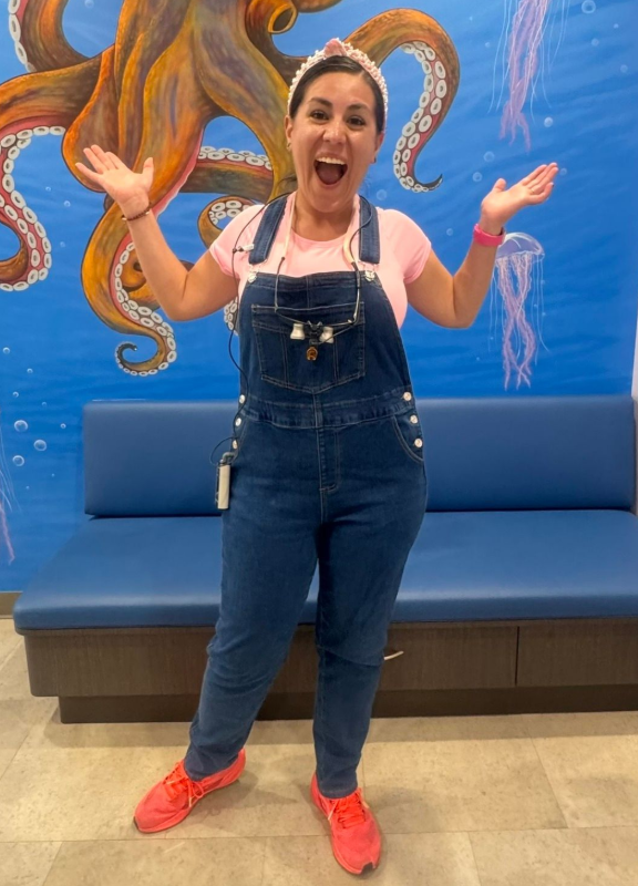 Woman posing with arms raised in front of a blue couch and octopus mural, wearing denim overalls and red shoes
