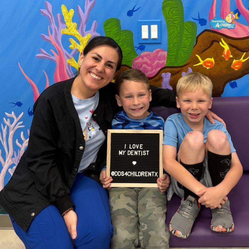 Smiling woman and two children posing with a “We love our dentist” sign in front of a colorful mural