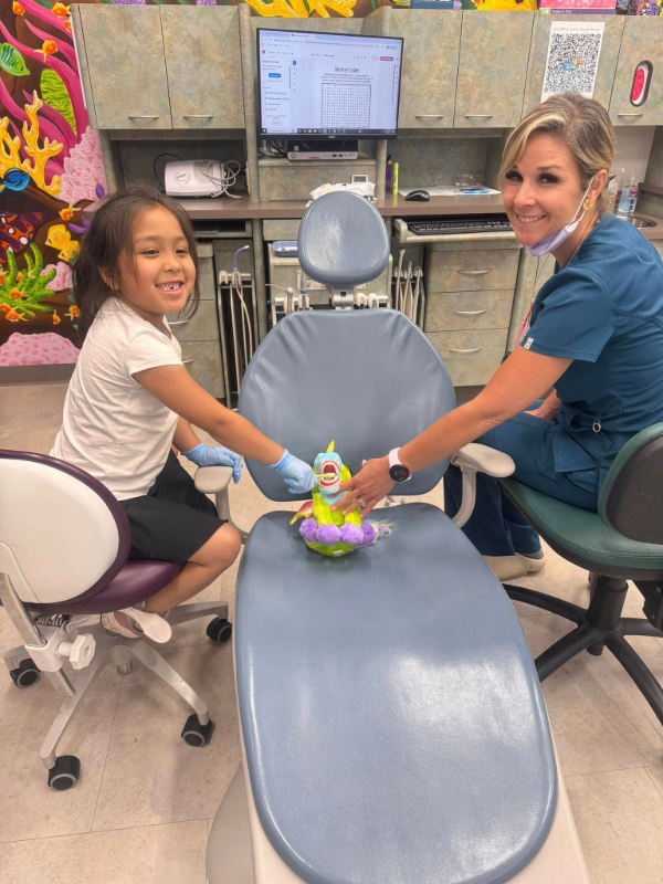 Dentist and young patient smiling beside a blue dental chair in a clinic, holding a toy on the chair.