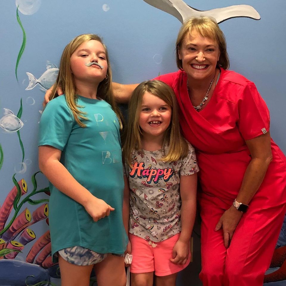 A woman in red scrubs is posing for a picture with two little girls one of whom is wearing a shirt that says happy