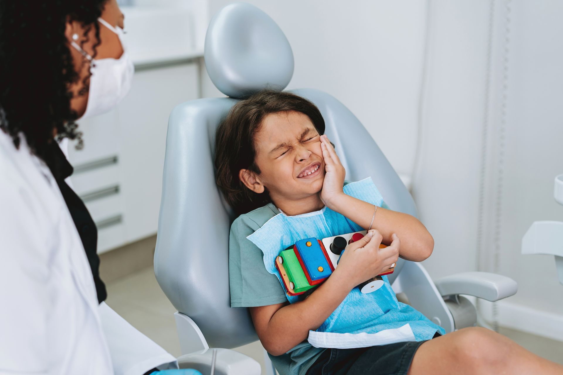 Boy in dentist chair holds cheek in pain as dentist in mask looks on.