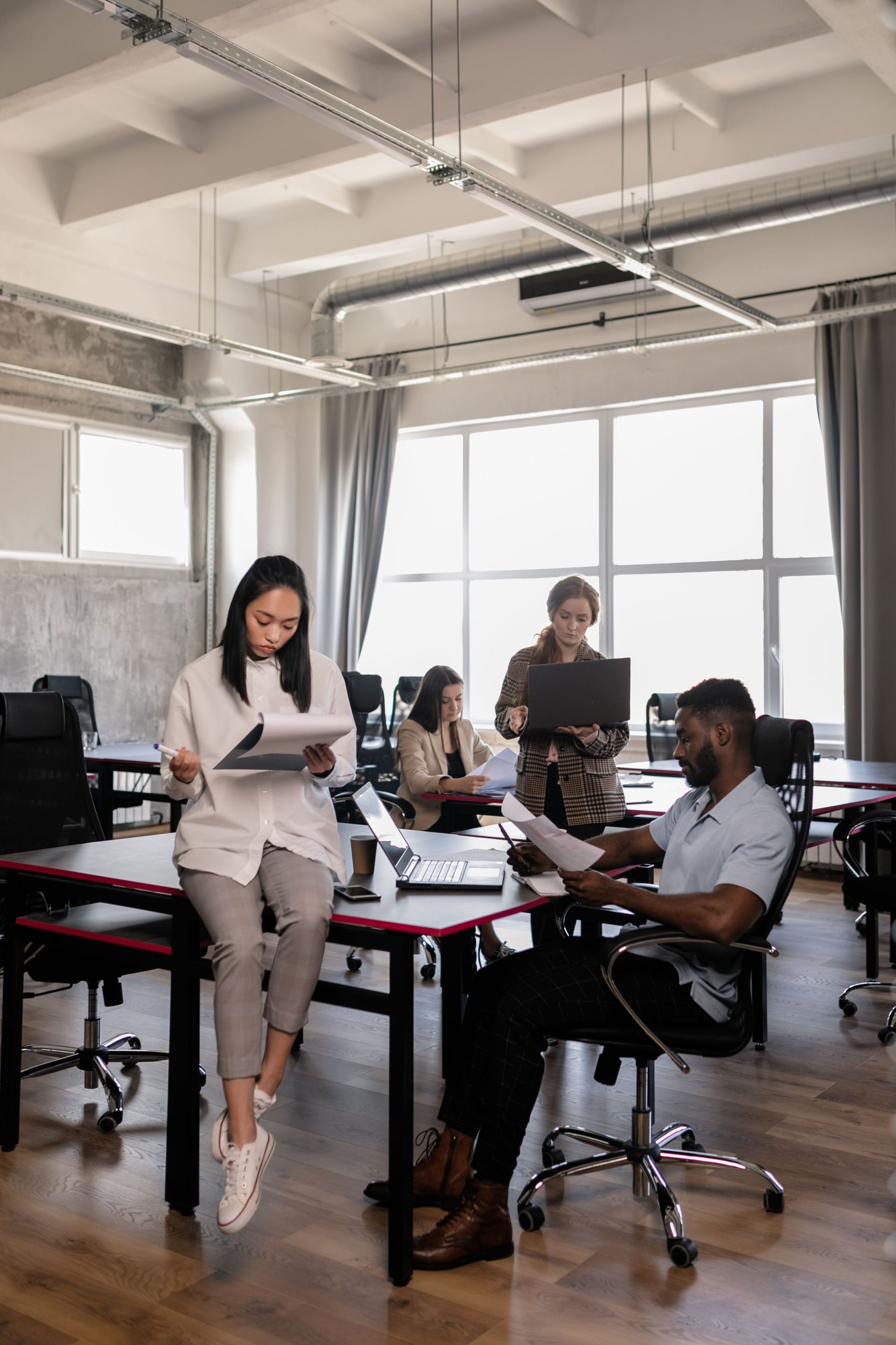 Personas colaborando en una oficina luminosa con escritorios, computadoras portátiles y grandes ventanas.