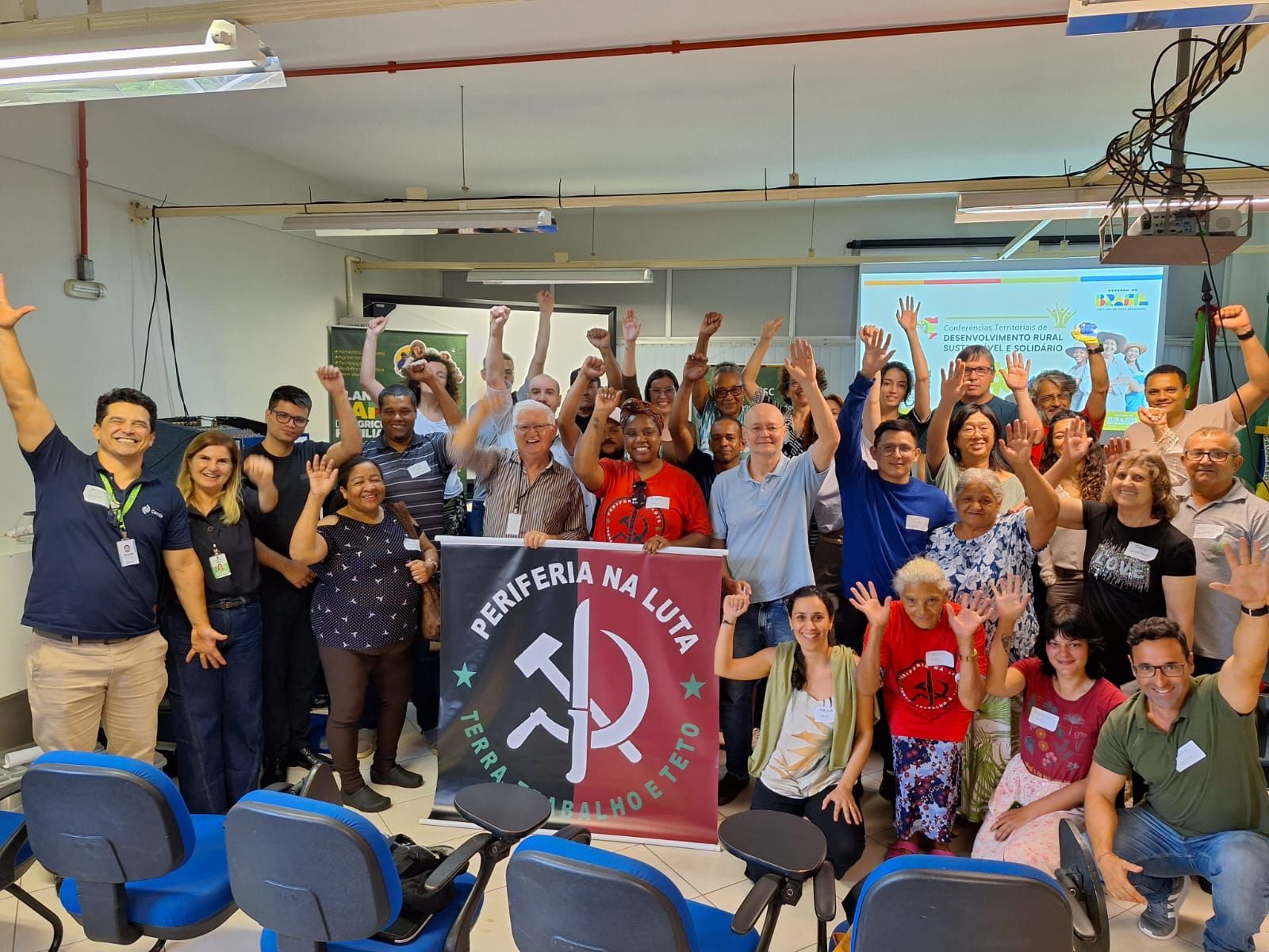 Grupo de pessoas sorridentes comemorando em ambiente fechado, com as mãos erguidas ao redor de uma faixa e um pódio.