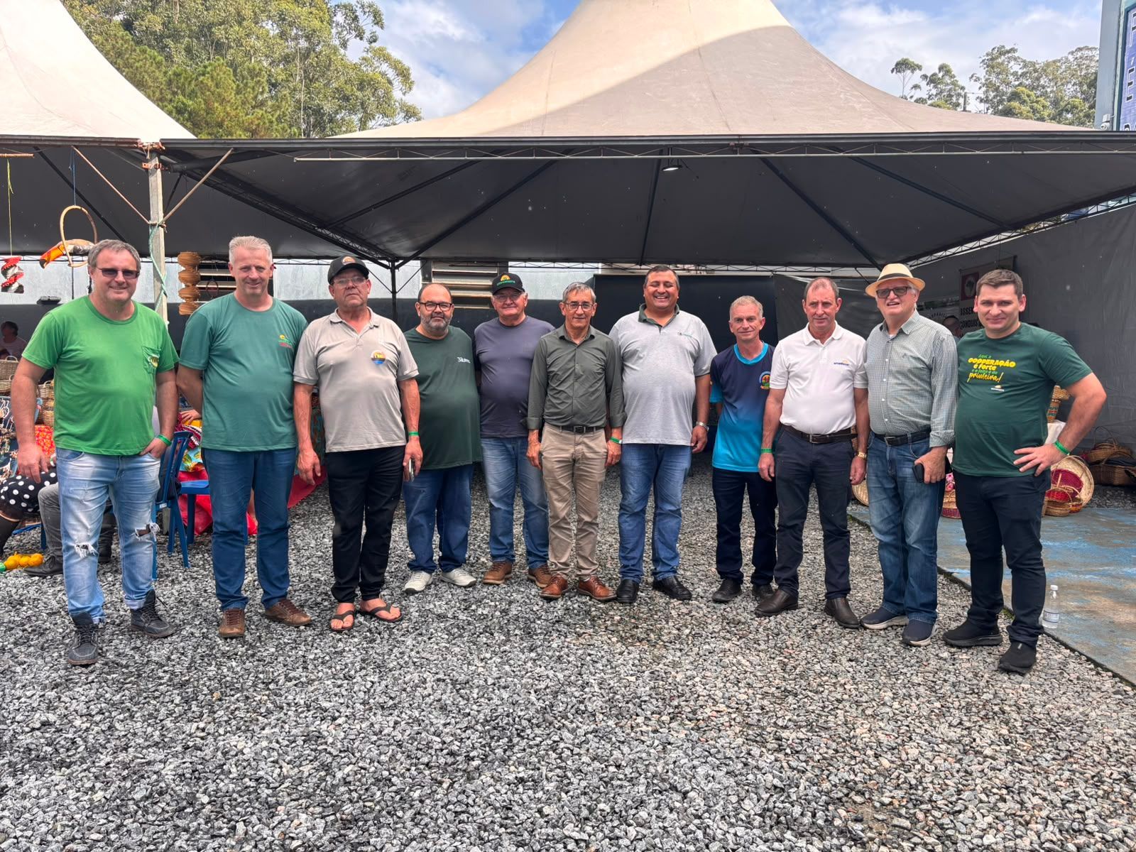 Grupo de homens em fila ao ar livre, sob um toldo preto sobre cascalho, com tendas e árvores atrás deles.