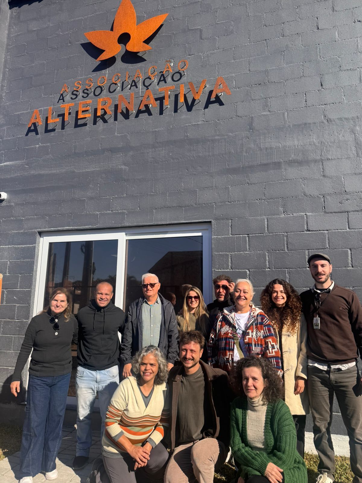Group of people posing outside a gray building with an orange “Associação Alternativa” sign.