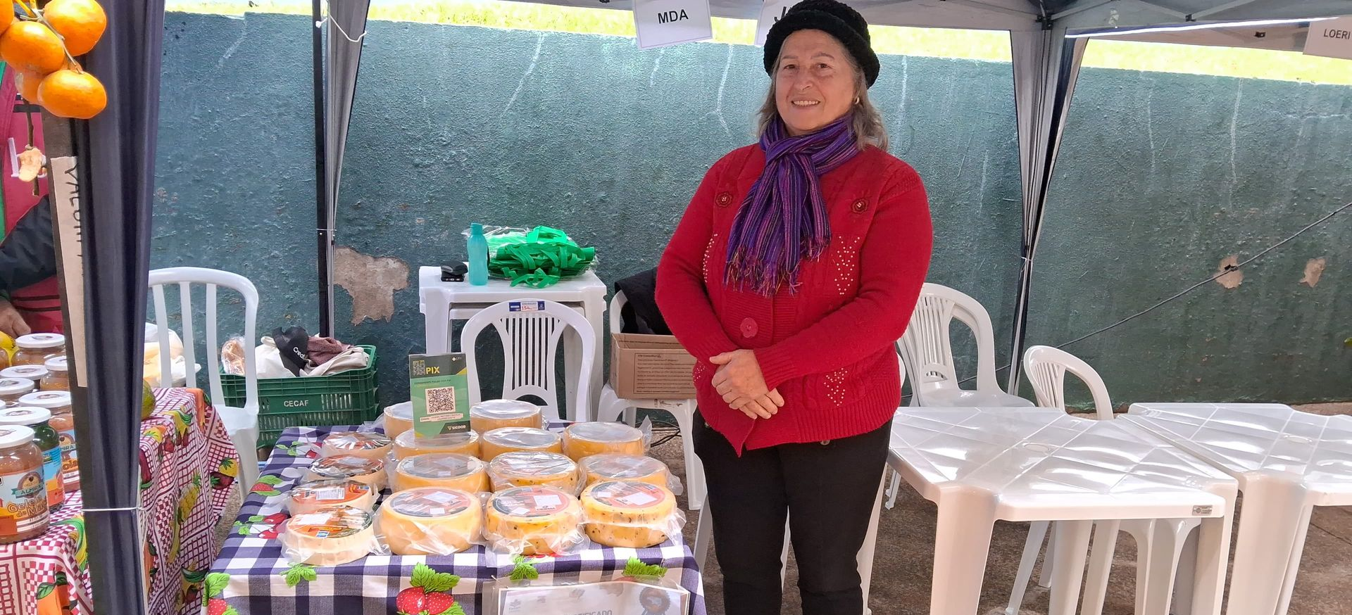 Pessoa em pé ao lado de uma mesa com alimentos embalados e cadeiras empilhadas em uma barraca coberta de mercado ao ar livre.