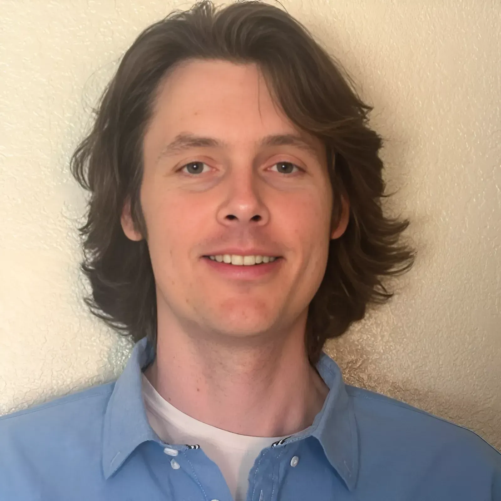 Portrait of a smiling person with shoulder-length dark hair wearing a light blue shirt against a beige wall