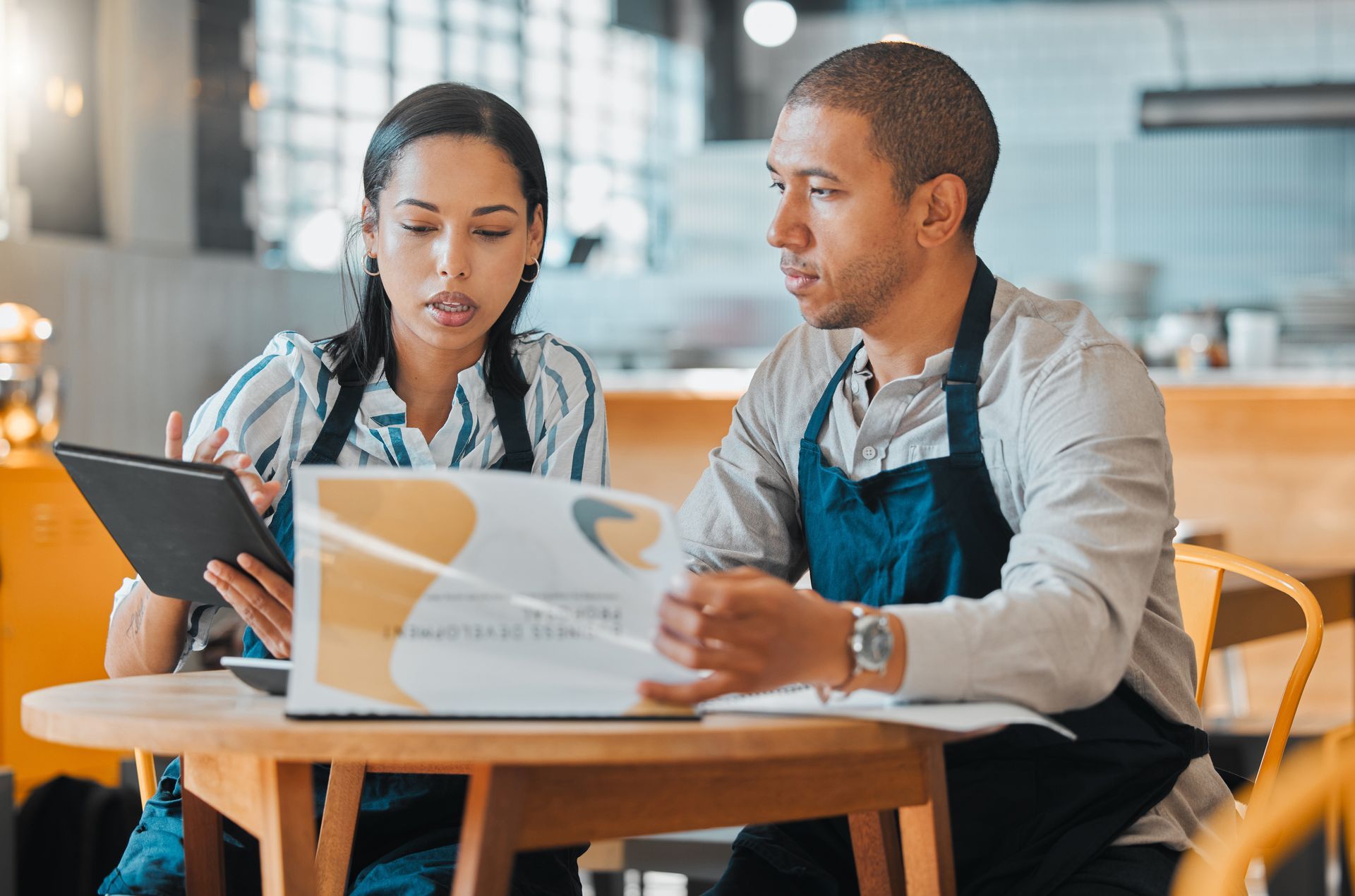 Two people in aprons reviewing documents at a table in a cafe, one holding a tablet, other holding paper.