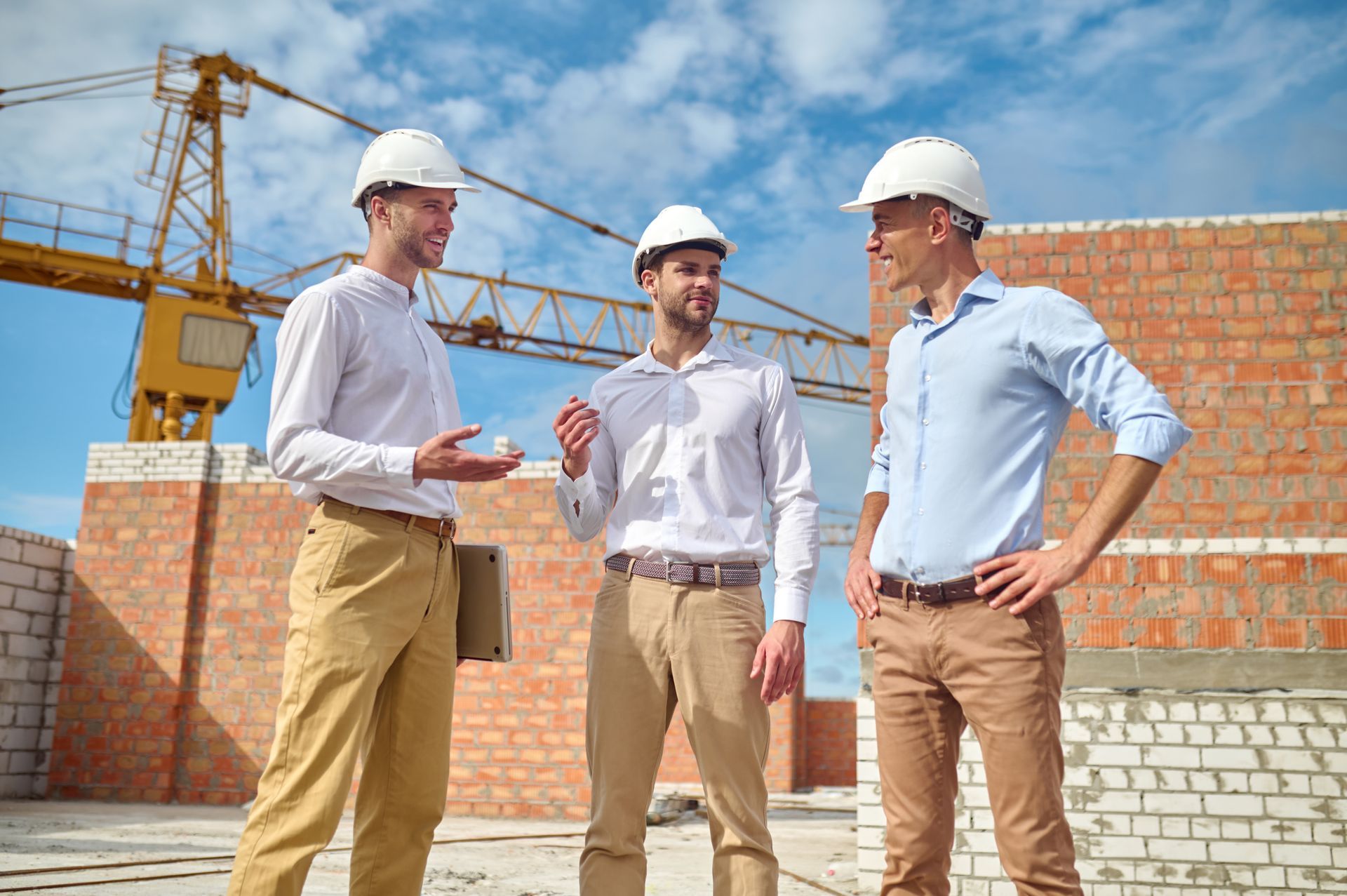 Three construction workers in hard hats discussing a project at a building site, crane in the background.