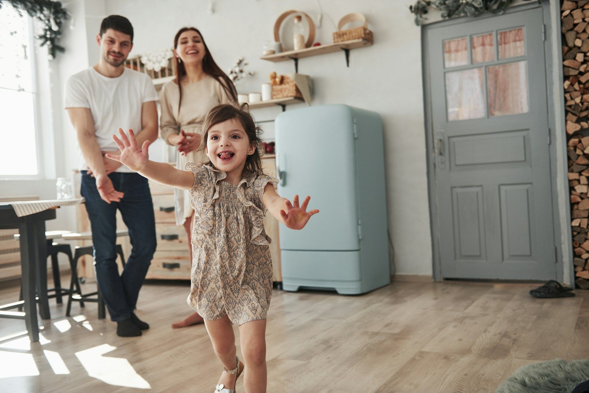 Smiling family in a bright kitchen, child running toward camera, parents standing behind near a blue fridge