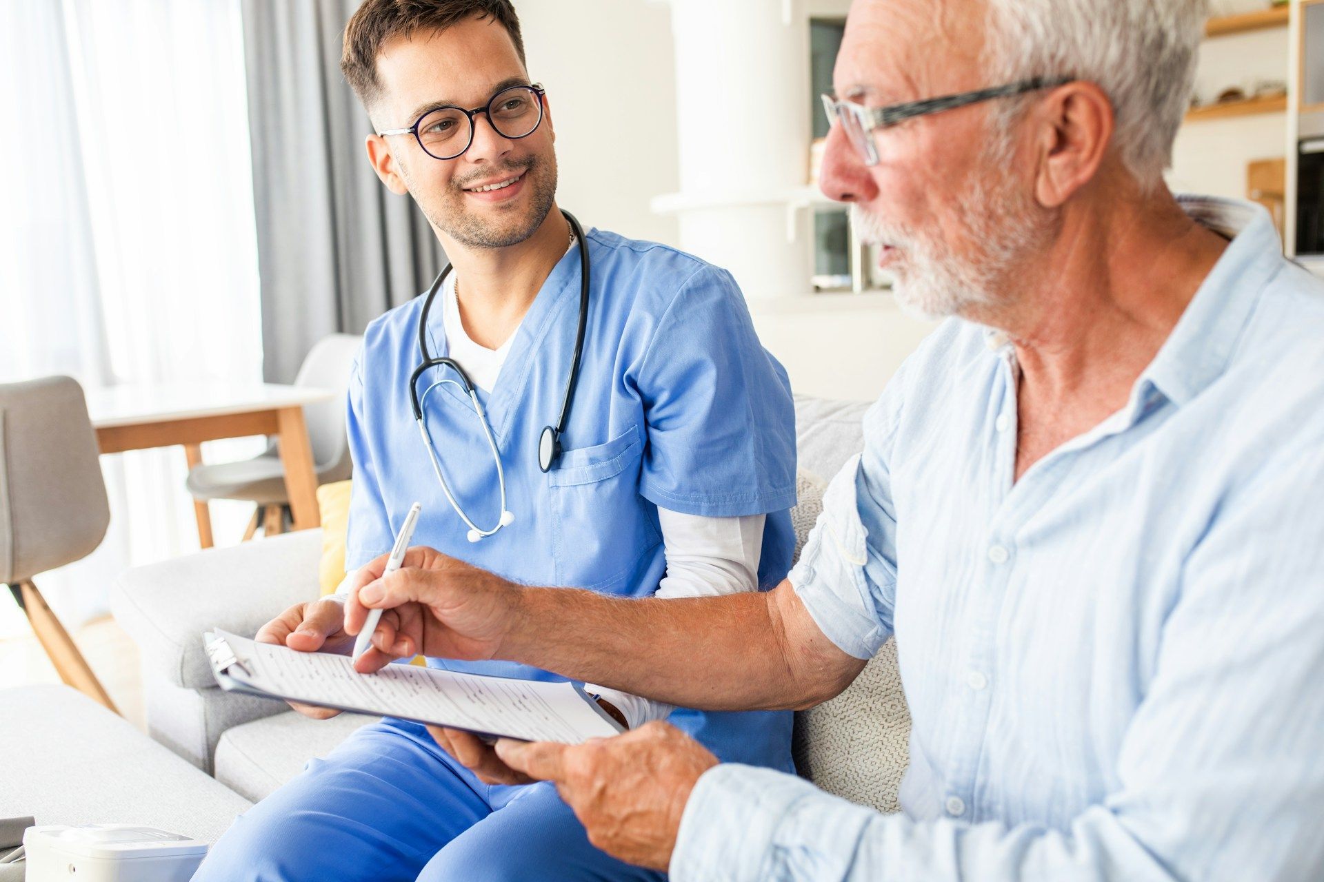 Nurse in blue scrubs talks with an older patient holding a clipboard in a bright clinic room