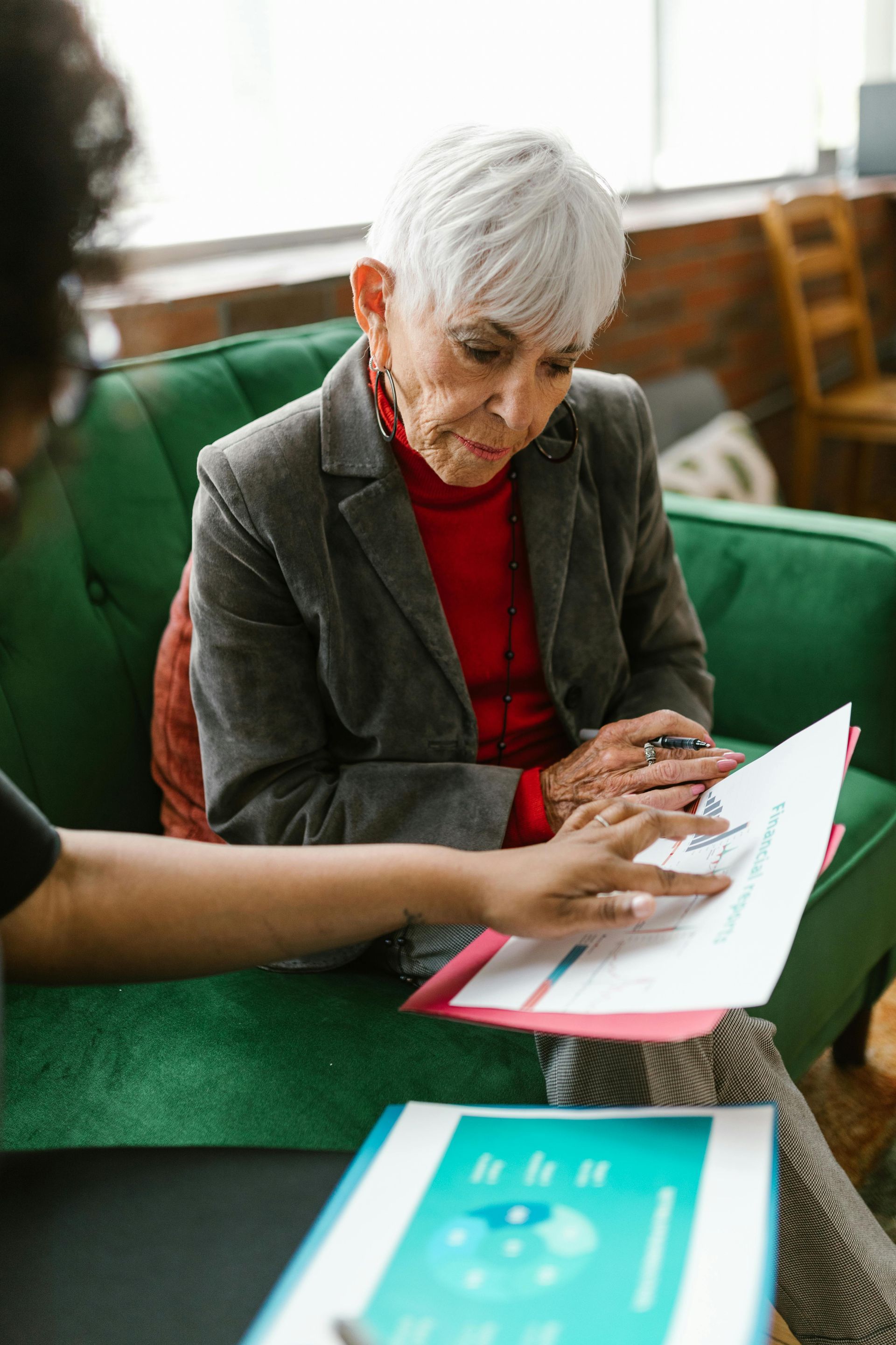 Older adult in gray jacket reviews documents with another person in a green sofa seating area