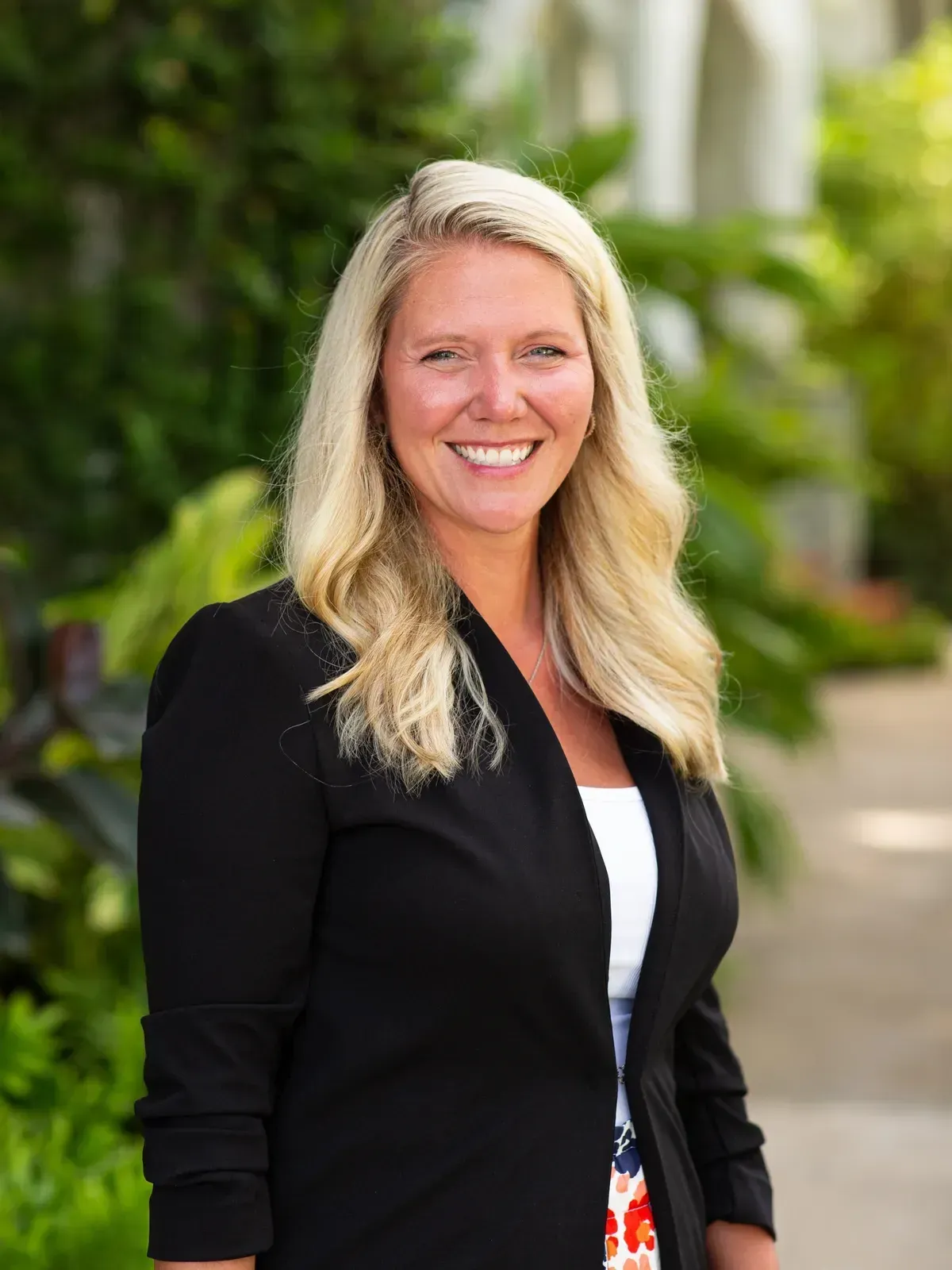 Smiling blonde woman in a black blazer standing outdoors with greenery in the background