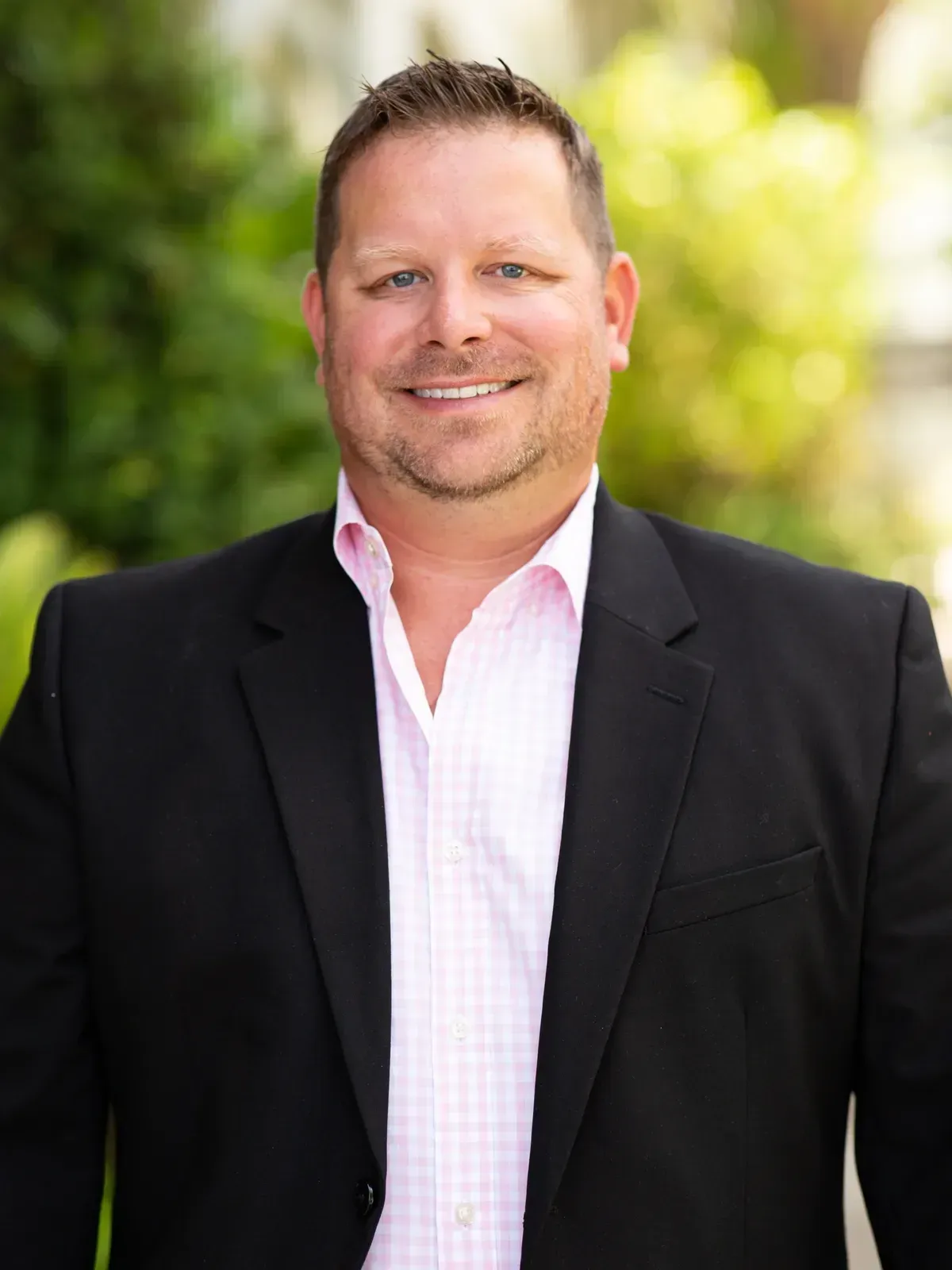 Smiling man in a black blazer and pink shirt standing outdoors with blurred greenery behind him