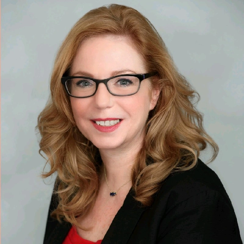 Woman with wavy blonde hair and glasses smiles. Wearing a red top and black jacket.