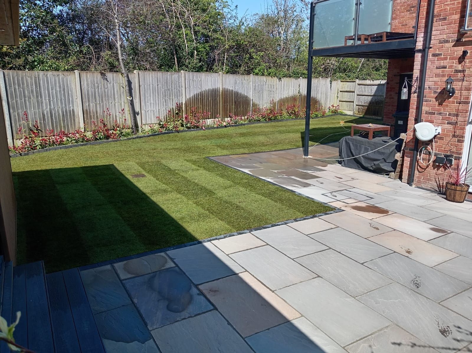 Backyard patio with gray paving, green lawn, fence, and brick house wall in sunlight