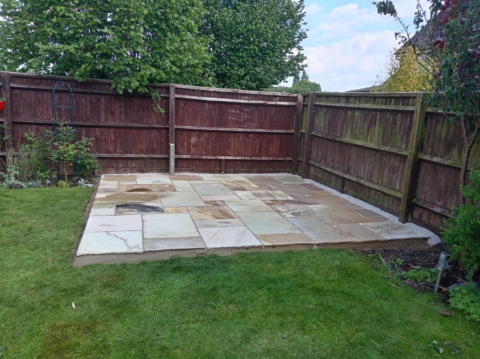 Backyard patio with stone pavers and wooden fence, bordered by grass and trees