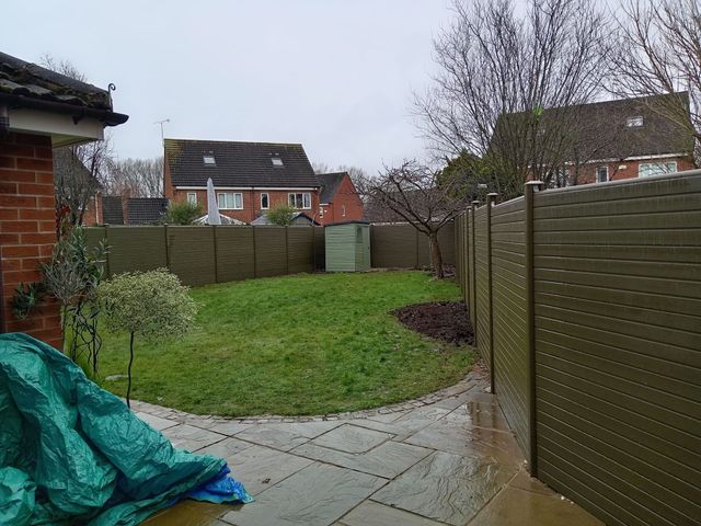 Backyard patio with green lawn, wooden fence, and houses beyond on a cloudy day