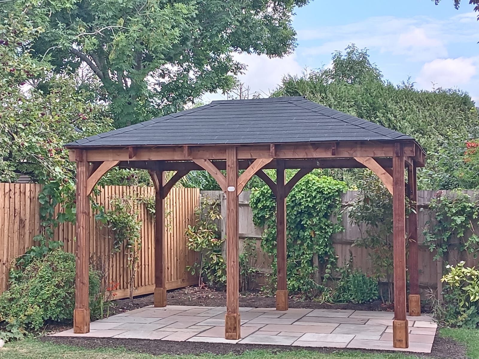 Wooden gazebo with a dark roof in a garden, surrounded by trees and a fenced patio.