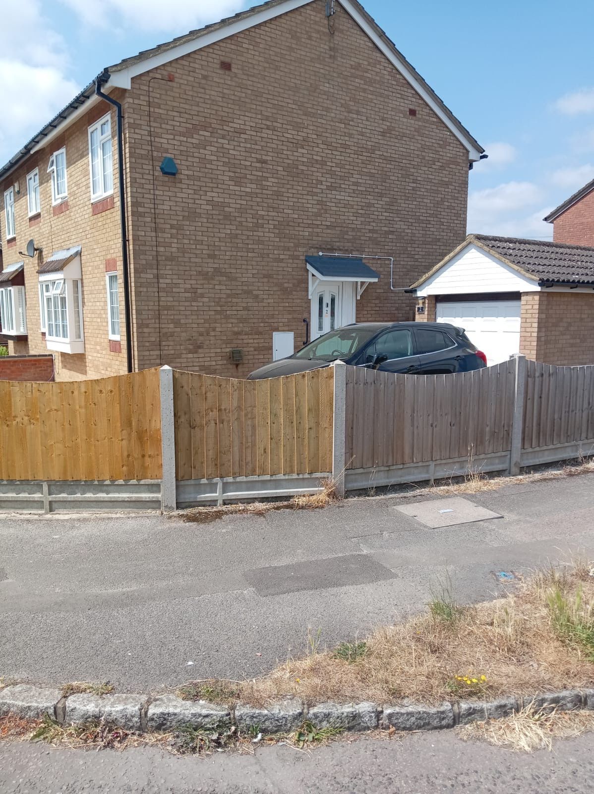 Residential street with a brick house, wooden fence, parked car, and overgrown curbside grass.