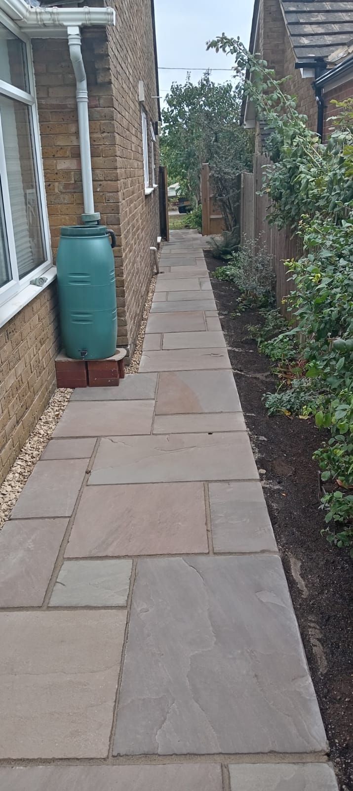 Narrow paved walkway between brick houses, with a green water butt on the left and hedges on the right