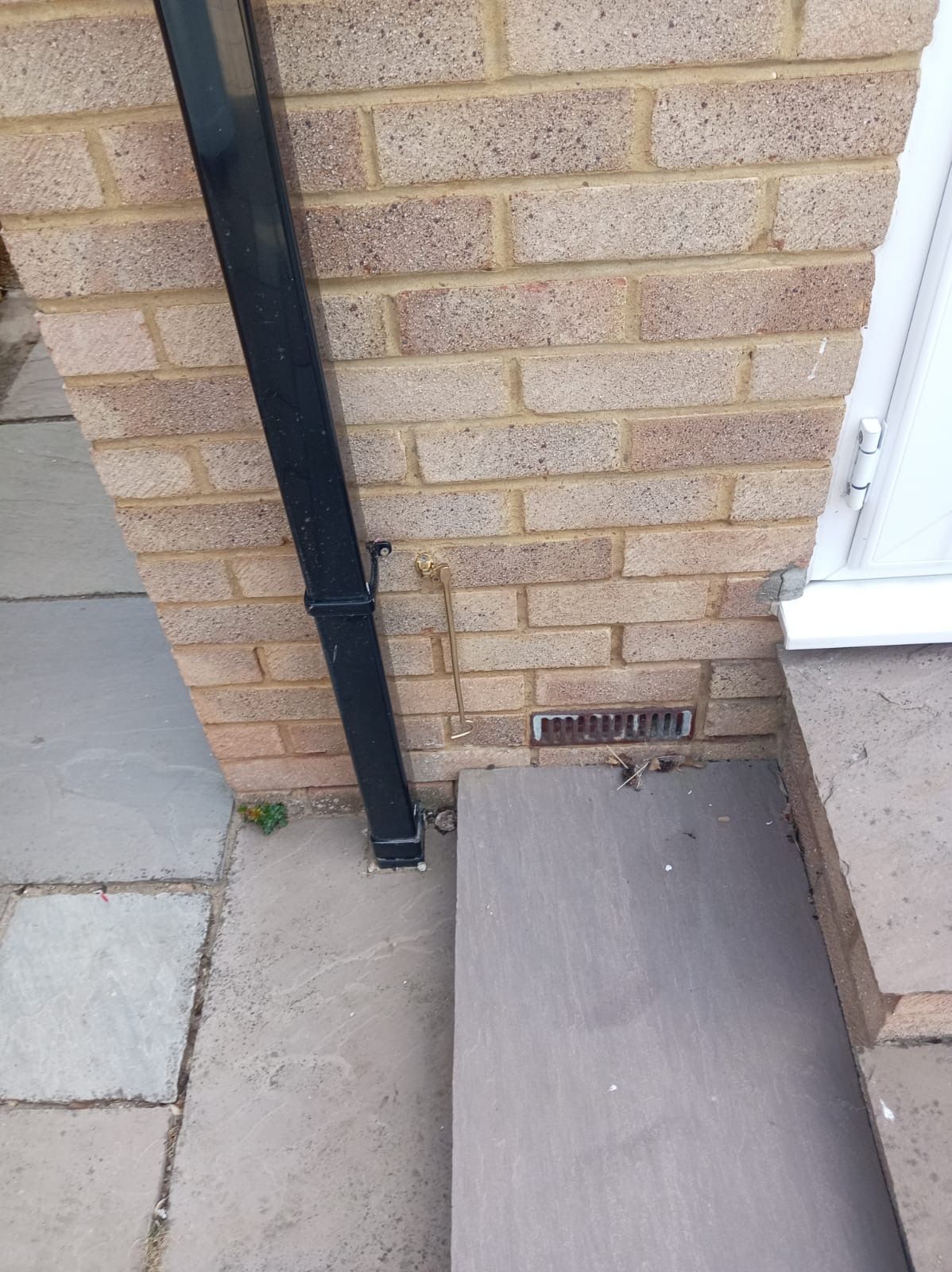 Brick house wall with black downspout, utility vent, and a gray ramp beside a paved path