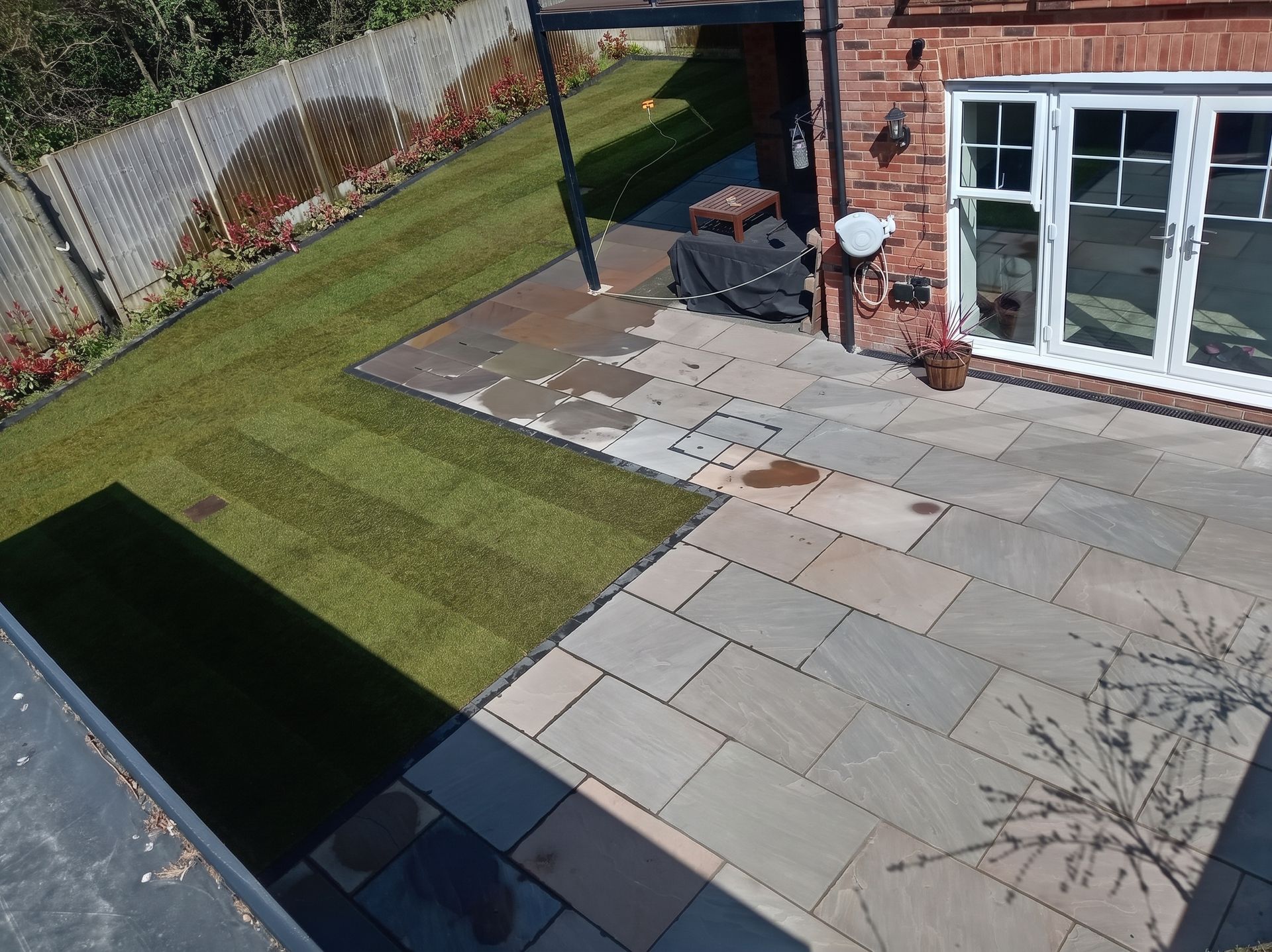Backyard patio with stone paving, sloped artificial grass, and a brick house wall with glass doors.