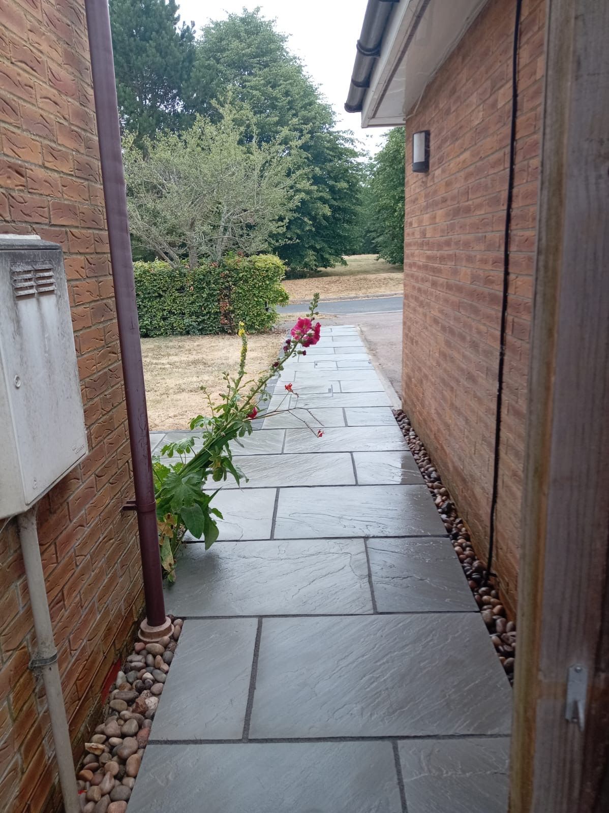 Narrow stone walkway between brick houses leading to a garden, with a red toy or bucket near the end.