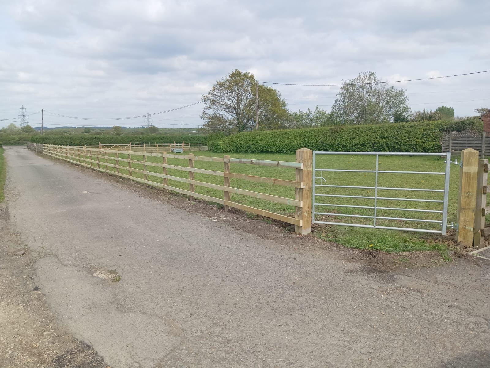 Rural roadside with a fenced green field and metal gate under a cloudy sky