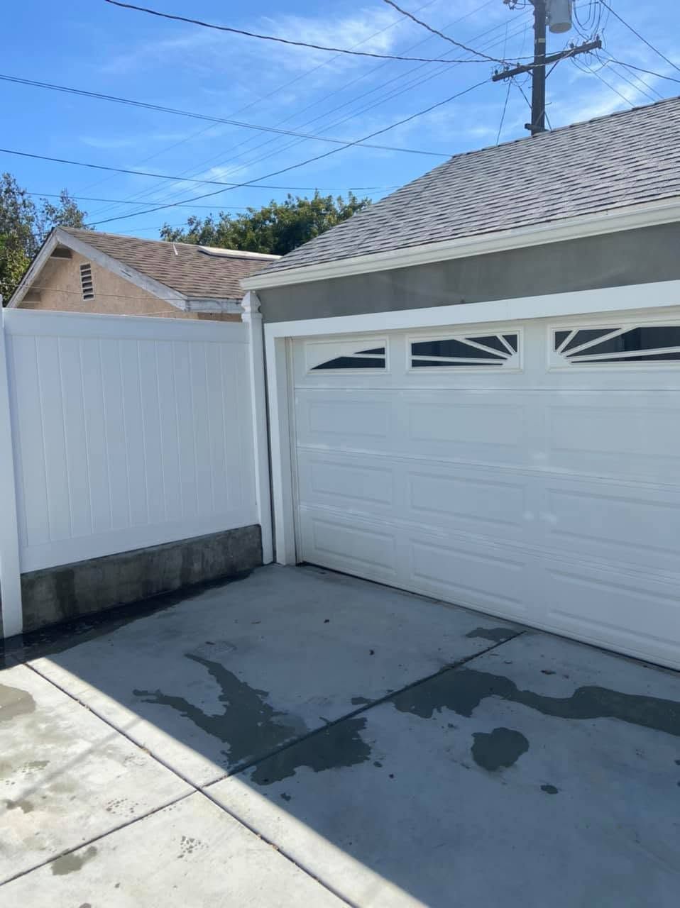 White garage door beside a white fence, with wet concrete driveway under a clear blue sky.