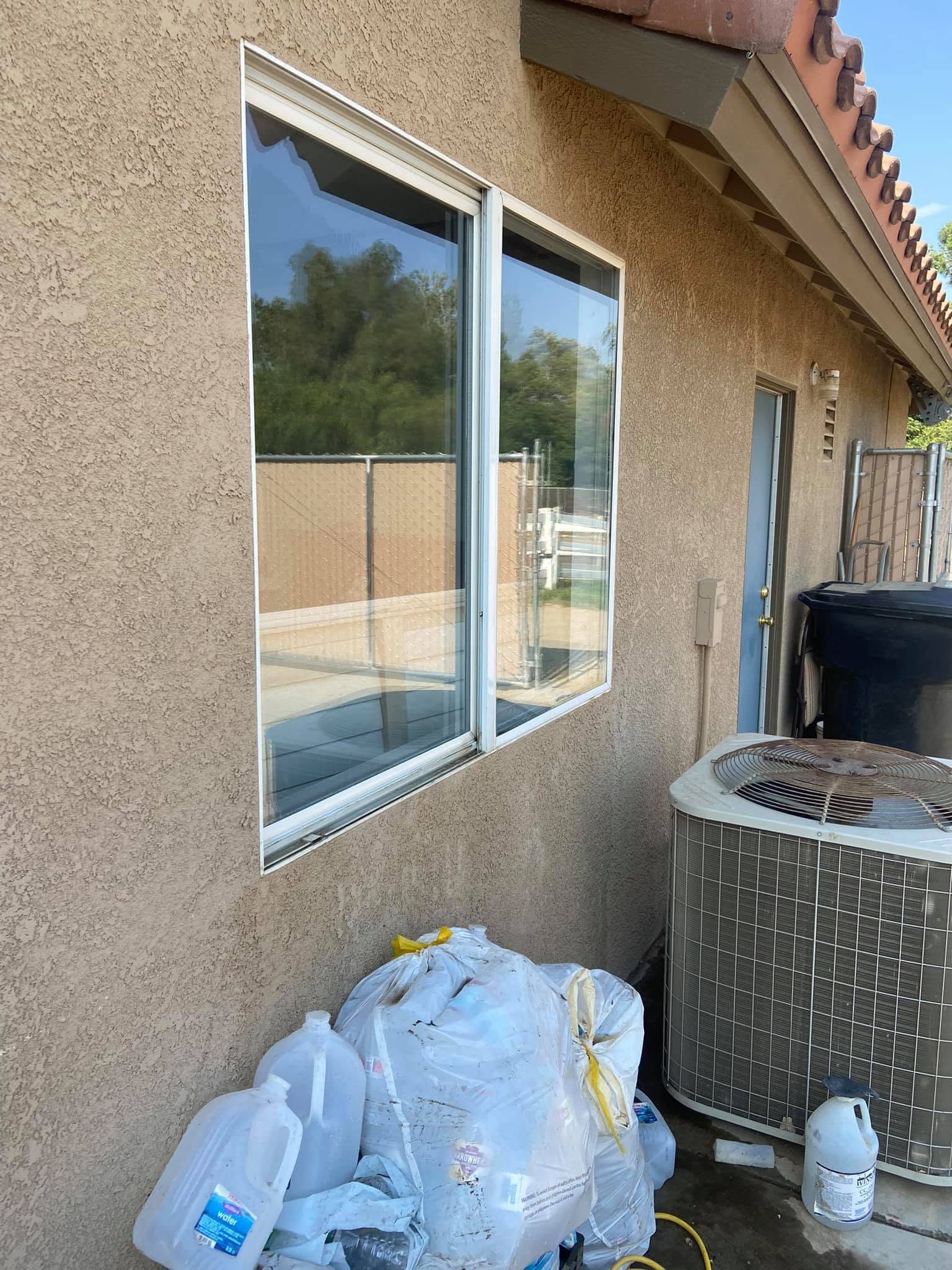 Side of a stucco house with a window, trash bags, and a large air conditioner unit near a patio area