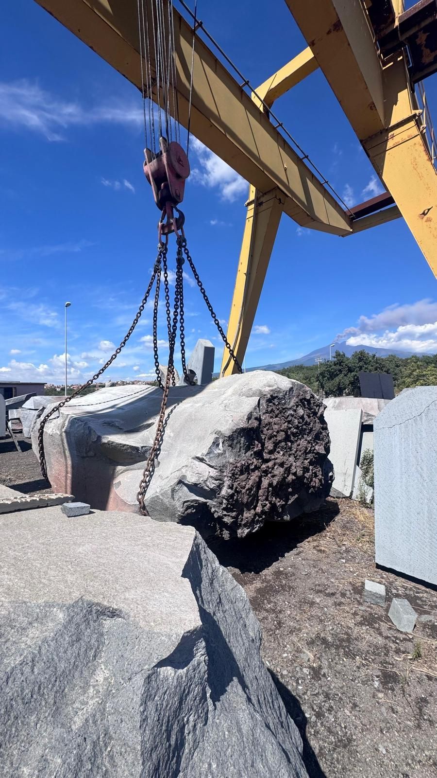 Grande roccia sospesa a una gru sotto il braccio giallo di un escavatore, in un piazzale ghiaioso con montagne e cielo azzurro sullo sfondo