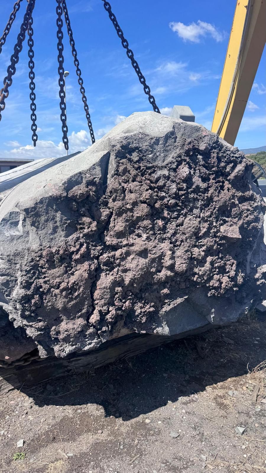 Grande masso grezzo grigio sollevato da catene di una gru contro il cielo azzurro