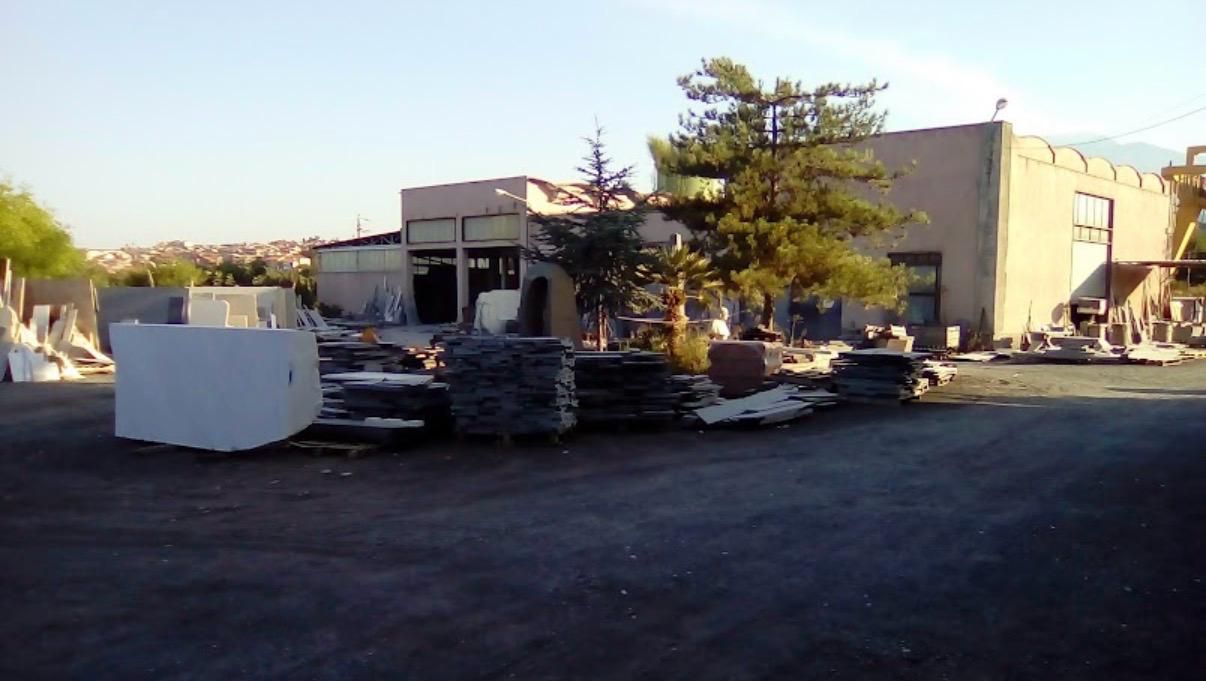 Piazzale industriale con materiali impilati, un container bianco e edifici beige in una giornata limpida.