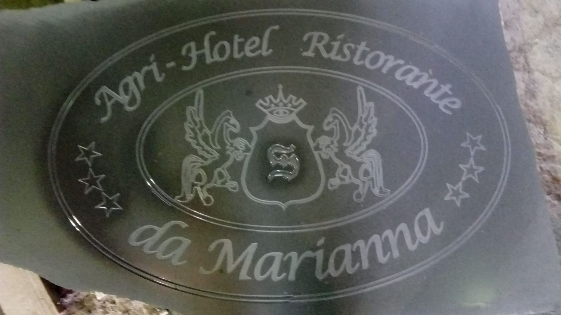 Insegna ovale di un hotel-ristorante con la scritta «Agri-Hotel Ristorante da Marianna» in bianco su sfondo scuro