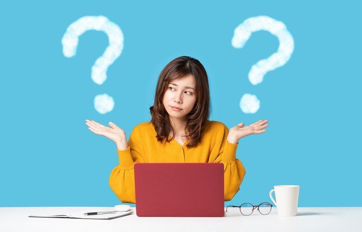 A woman is sitting at a desk with a laptop and two question marks above her head.