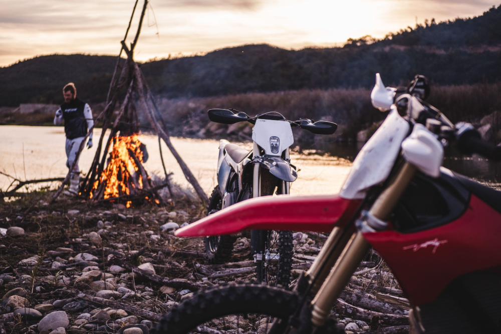 Two dirt bikes near a campfire on a lake shore. A person stands by the fire at dusk.— EVA Motors in Paget, QLD