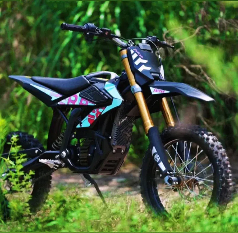 Black and turquoise electric dirt bike on grass, surrounded by greenery.
