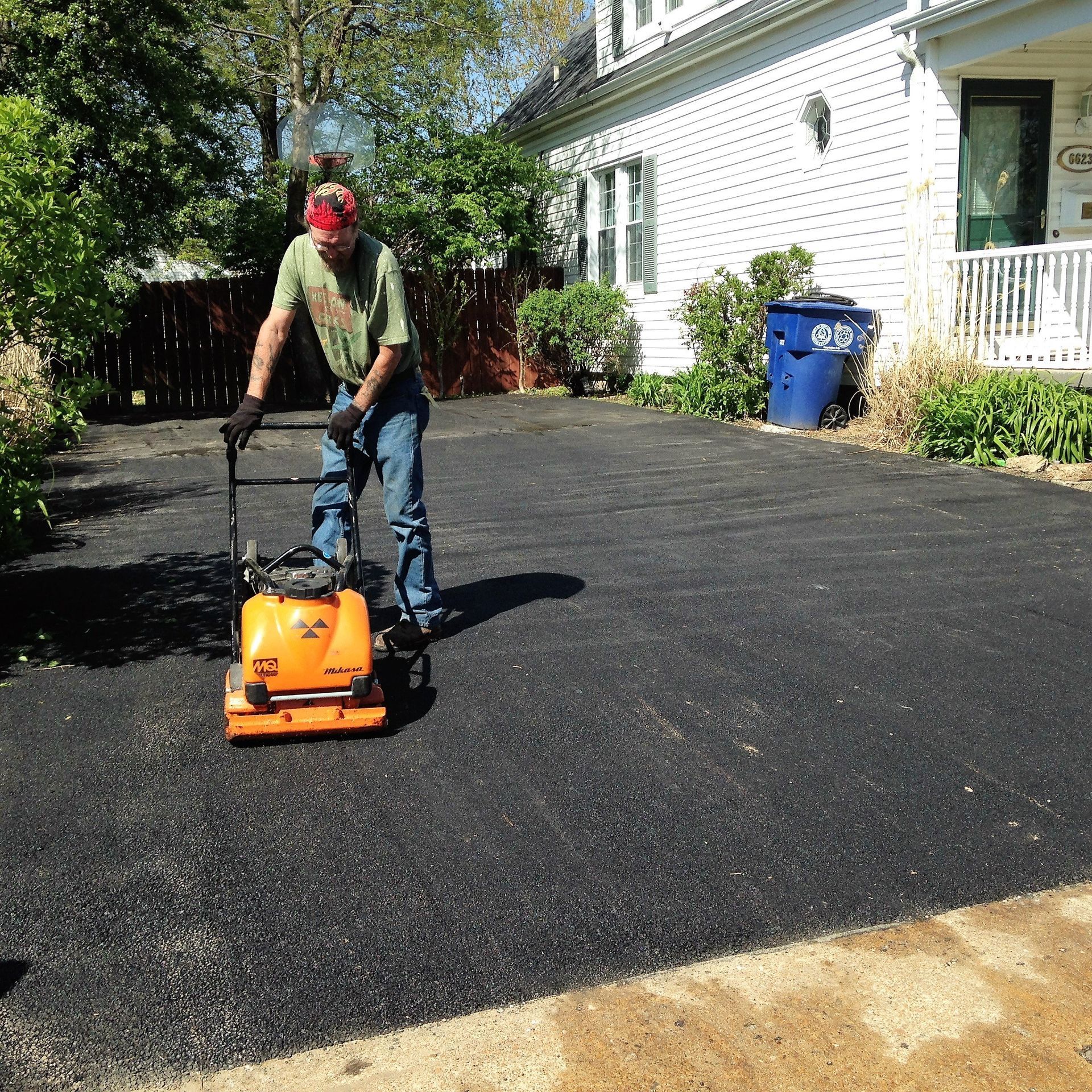 Worker operating an orange asphalt compactor on a driveway beside a house
