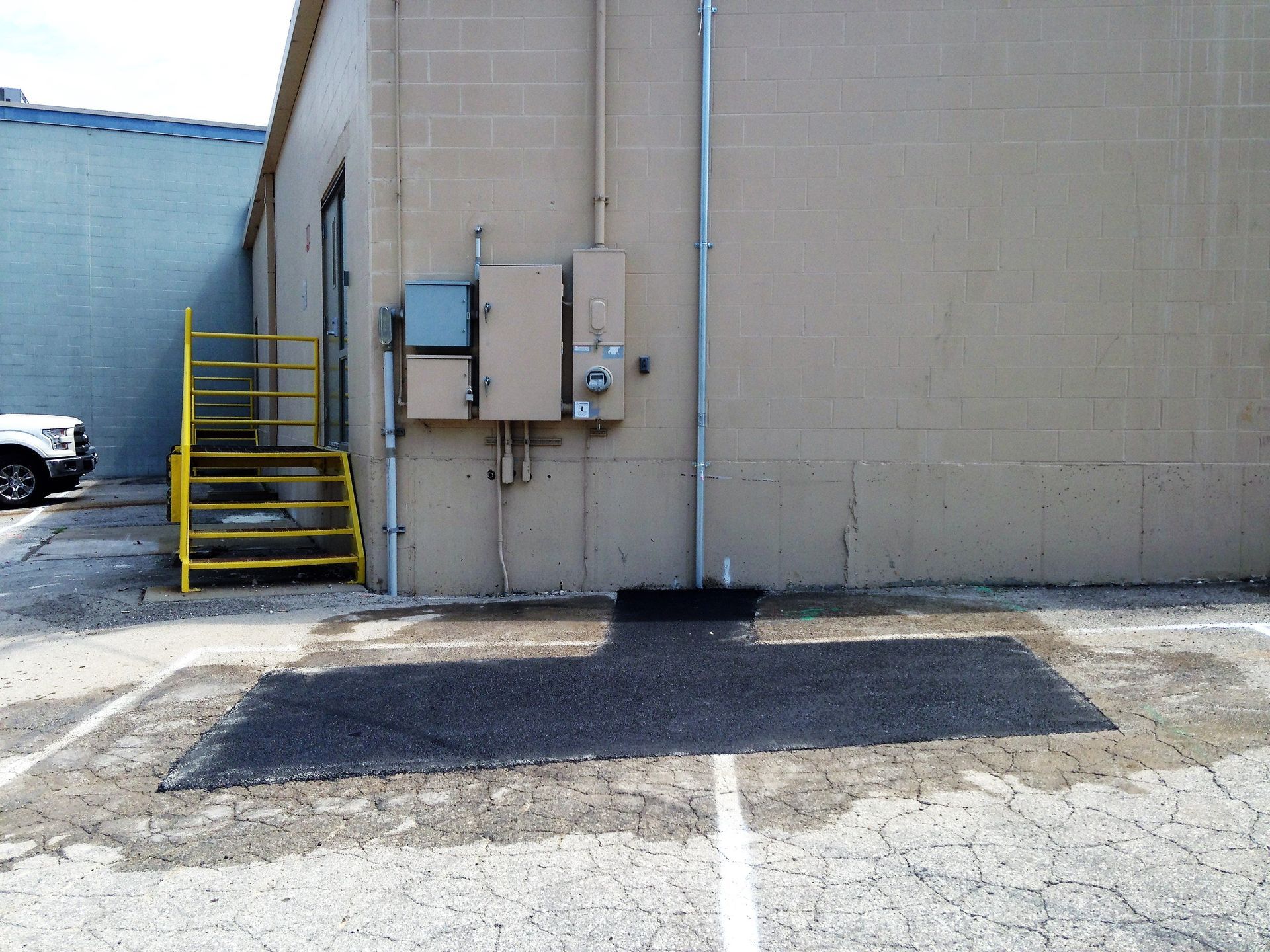 Alley beside a beige building with utility boxes, yellow stairs, and a cracked asphalt lot.