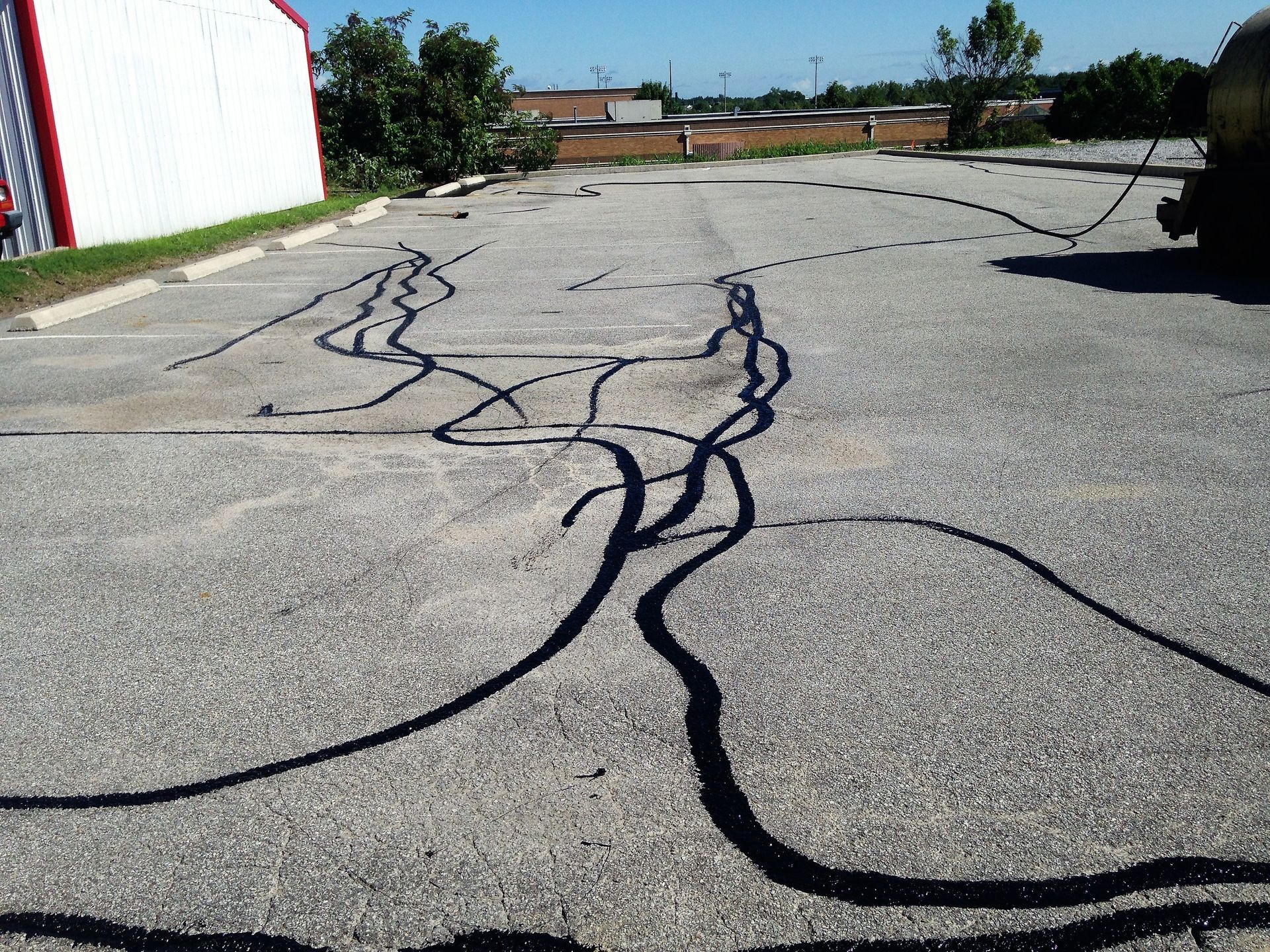 Cracked asphalt parking lot with long dark pavement cracks beside a white building and trees.