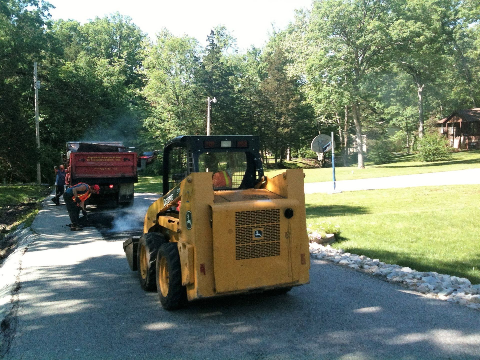 Yellow skid steer and red dump truck on a tree-lined residential road, with grass and homes nearby
