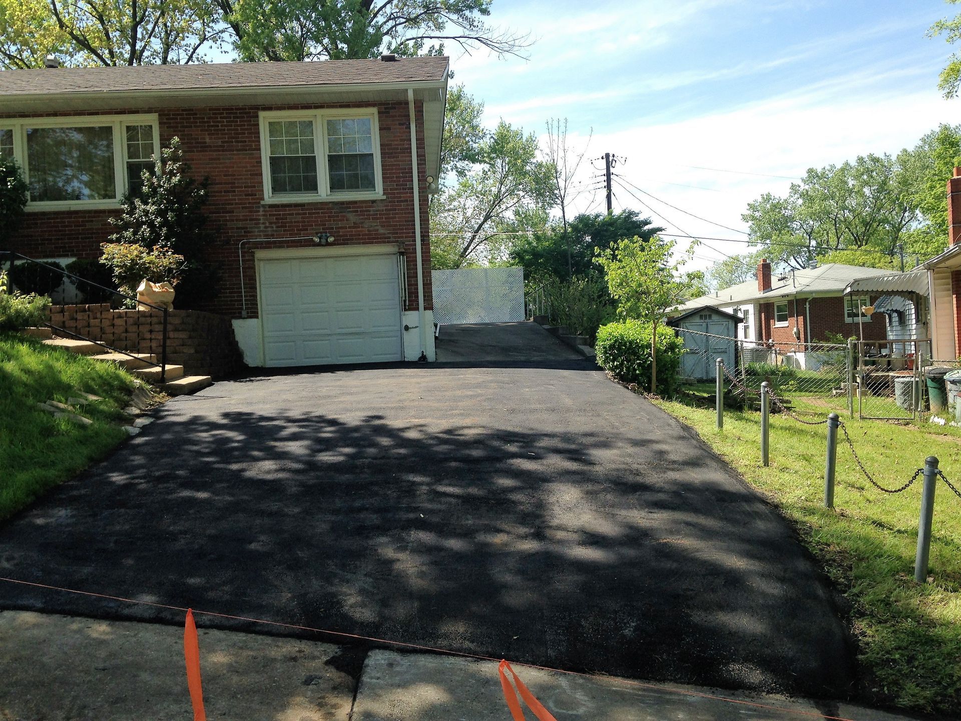 Residential driveway freshly paved beside a brick house, sloping toward a garage and street.