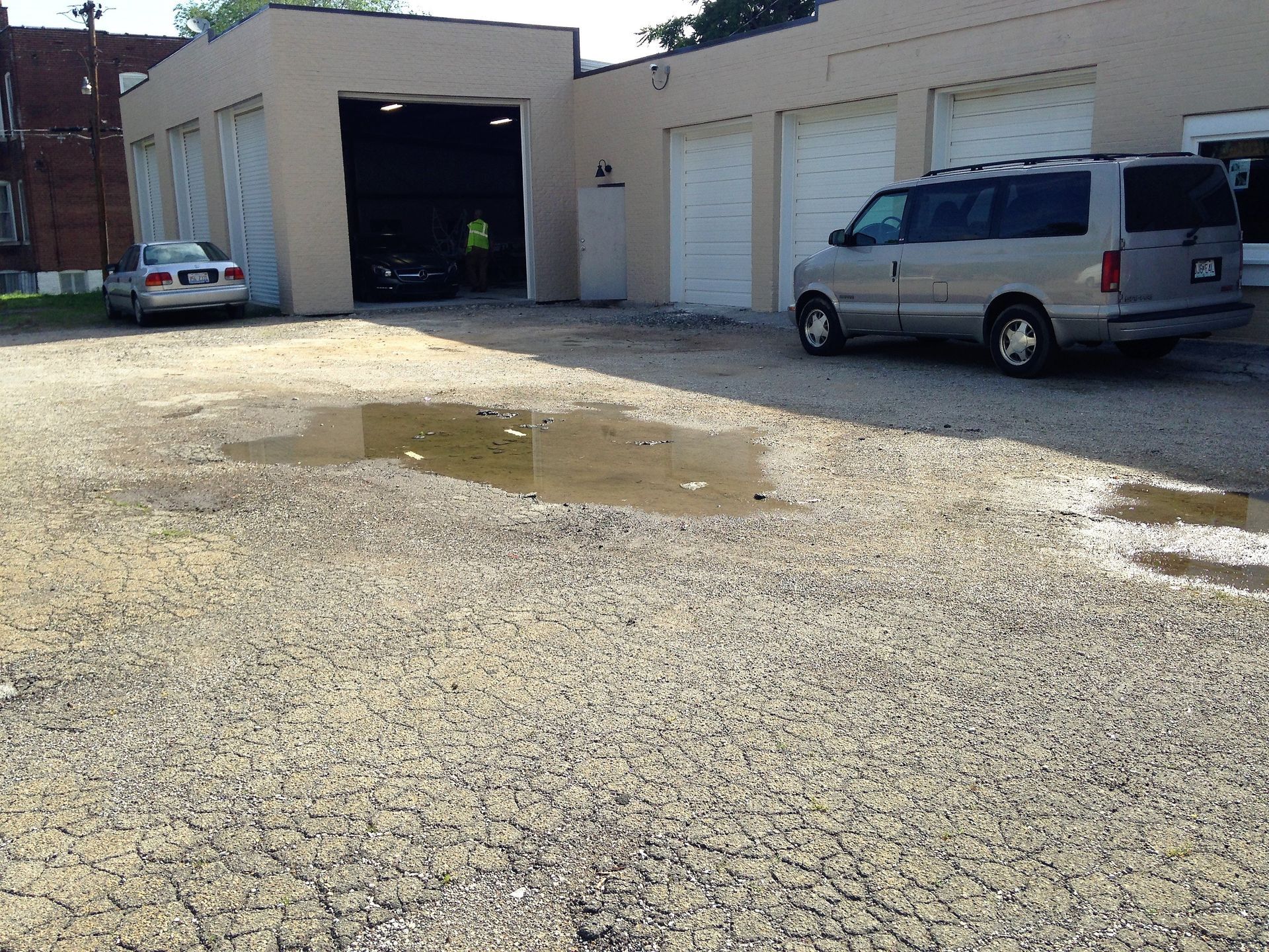 Industrial garage lot with puddles, a white van, and a blue car by open service bays.