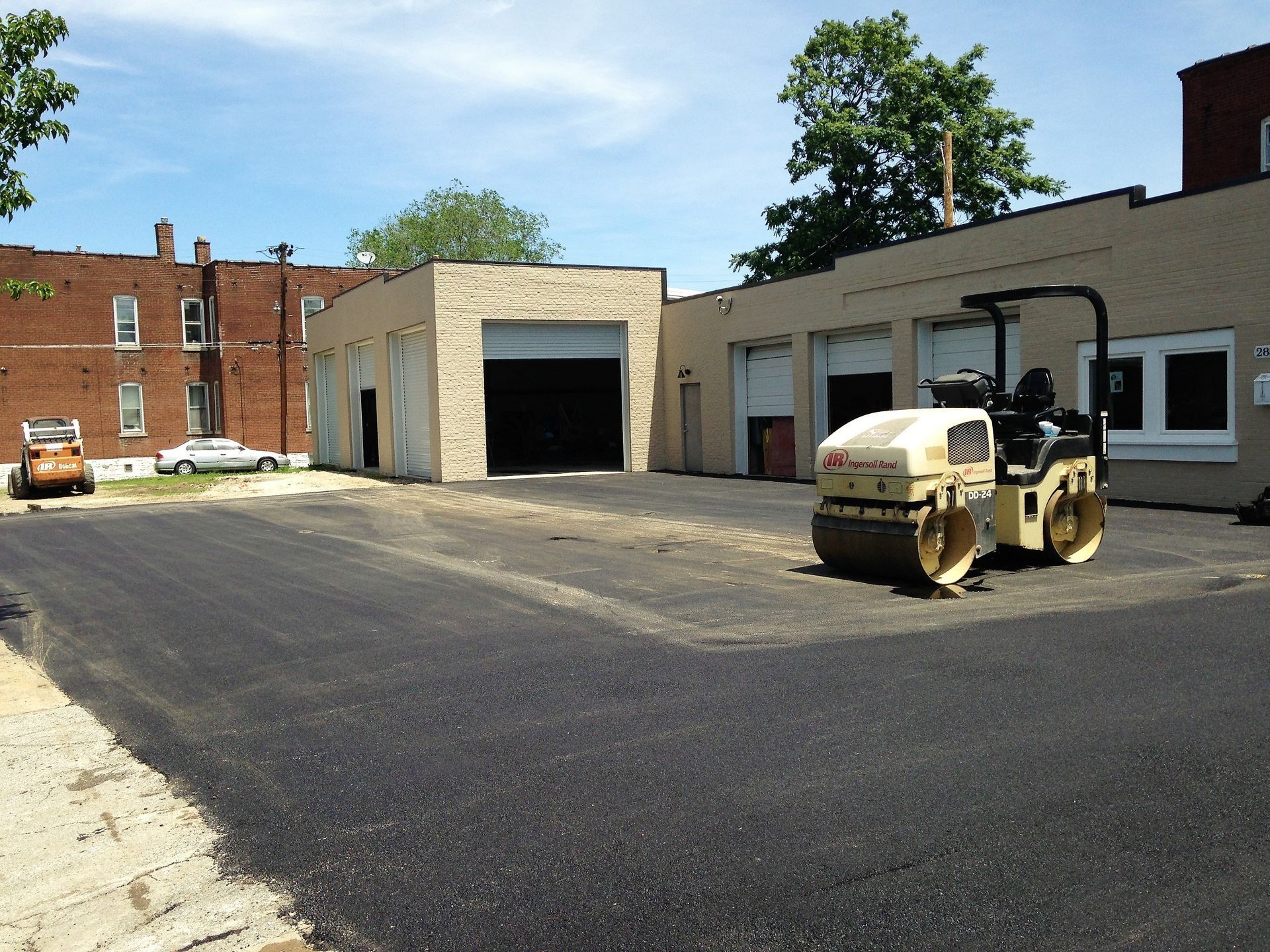 Road roller paving a freshly asphalted lot beside an industrial building on a sunny day