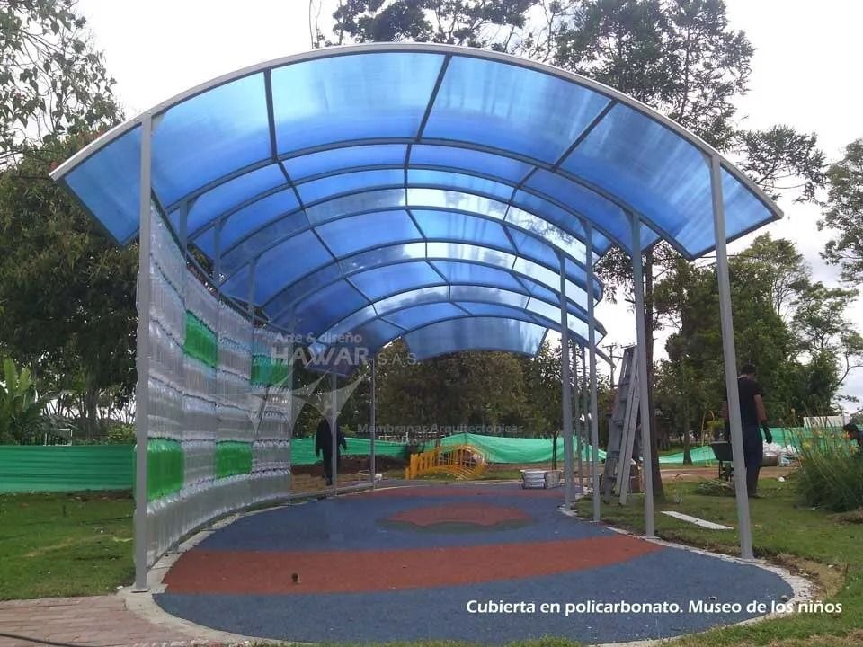 Pasarela cubierta con techo de policarbonato azul y estructura de acero en un sendero del parque.