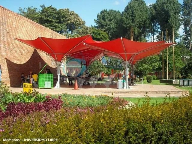 Patio exterior con toldos rojos, una entrada de cristal y plantas ornamentales bañadas por la luz del sol.
