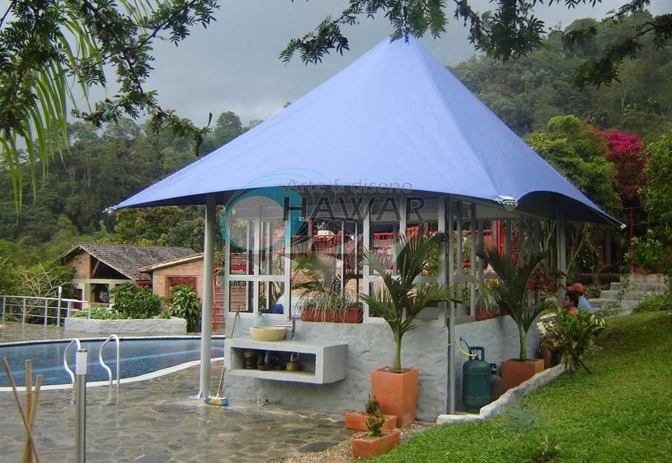 Pequeño cenador junto a la piscina con techo de lona azul en un entorno de jardín tropical.